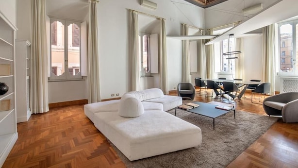 Un amplio salón con un sofá blanco grande, una mesa de centro y una mesa de comedor con sillas. Se ve la ciudad a través de amplios ventanales.