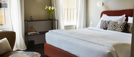 Una cama bien hecha con sábanas blancas, un cabecero rojo y almohadas estampadas. Hay una mesita redonda con un diseño colorido, una silla y una estantería con libros y un jarrón de flores.