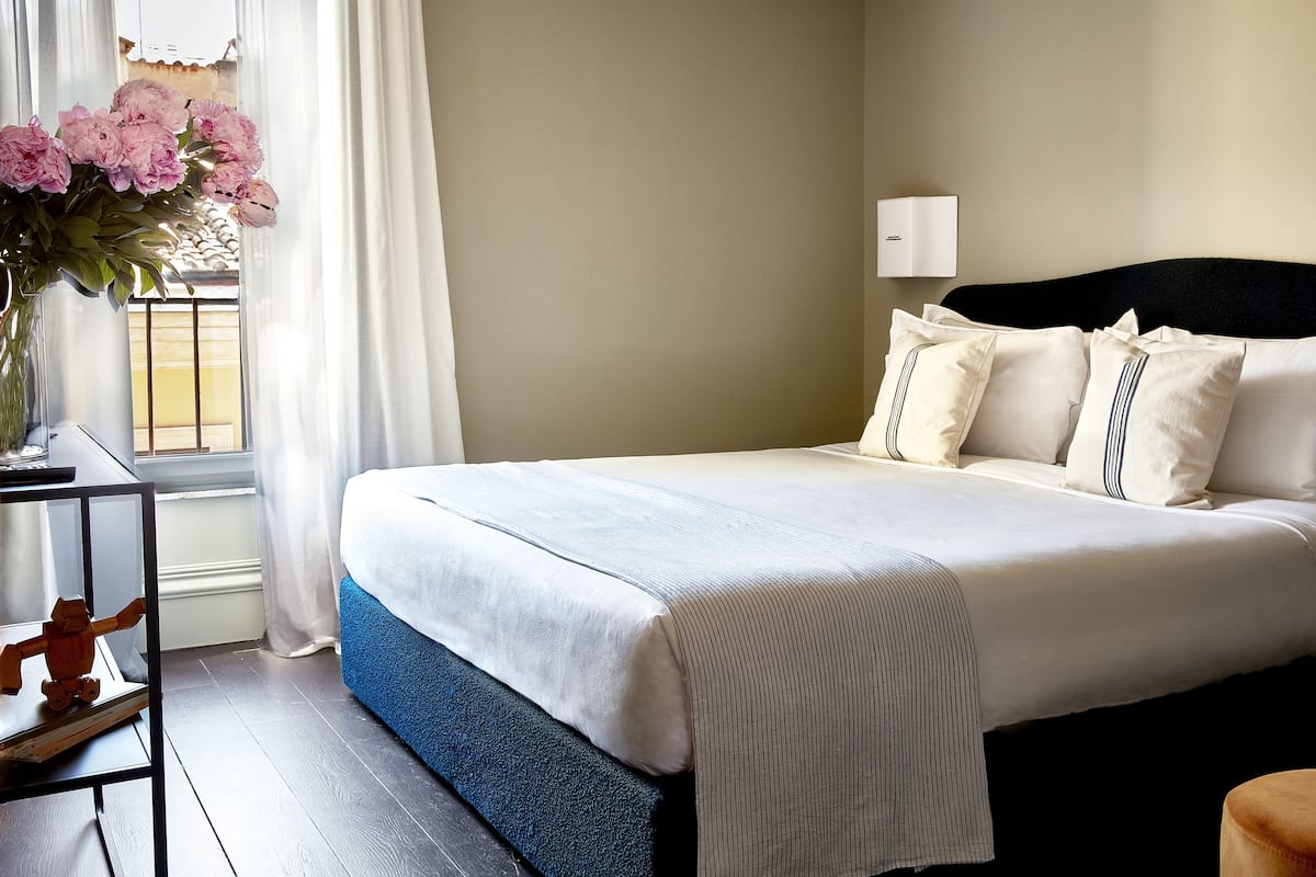 Una cama bien hecha con una colcha blanca y azul, una lámpara de noche y un jarrón con flores rosadas sobre una mesita de noche.