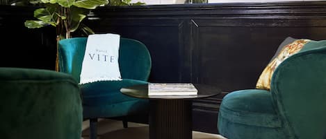 A teal upholstered chair with a 'Hotel VITE' placard, a small round table with a book, and a large green plant in a room with dark wooden paneling.