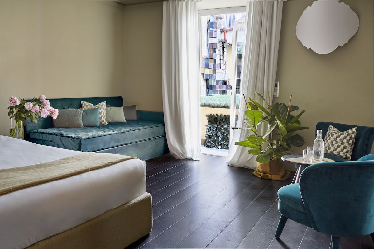Habitación de hotel con cama, sofá, sillón, una mesita con vasos y una botella, y vistas al exterior.