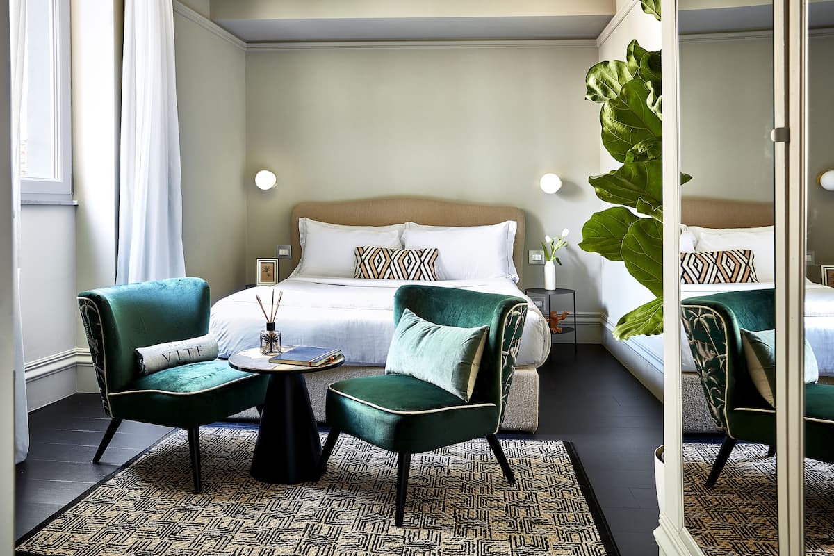 Habitación de hotel con una cama, dos sillones verdes, una mesita redonda y una planta grande.