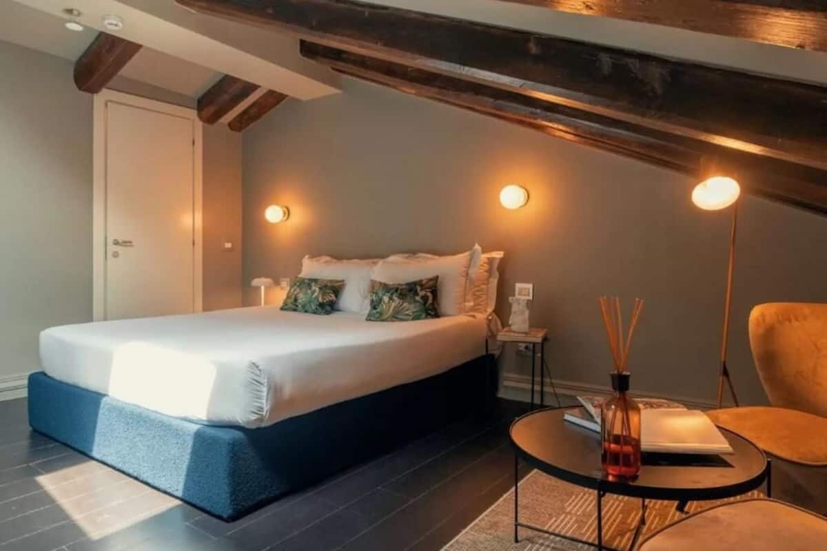 Un dormitorio con cama, una mesita de noche, una silla y una pequeña mesa con un libro y una botella.