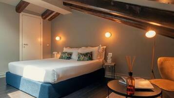 A bedroom with a bed, a bedside table, a chair, and a small table with a book and a bottle.