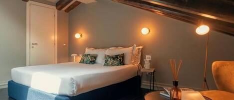 A bedroom with a bed, a bedside table, a chair, and a small table with a book and a bottle.