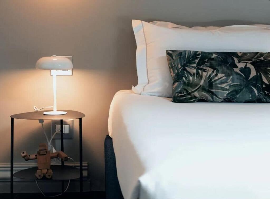 Un lit bien fait, avec une literie blanche et un coussin orné d’un motif de feuilles tropicales, une petite table ronde avec une lampe blanche et une figurine en bois posée sur la table.