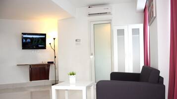 A modern living room with a flat-screen TV mounted on the wall, a small white table with a potted plant, a dark-colored sofa, and a door with frosted glass panels.
