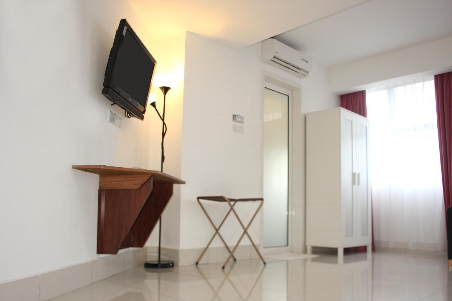 A hotel room with a flat-screen TV mounted on the wall, a wooden podium, a folding chair, a wardrobe, and an air conditioning unit.