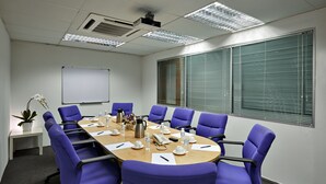 Uma sala de conferências com uma mesa comprida, cadeiras roxas, um quadro branco e um vaso de plantas.
