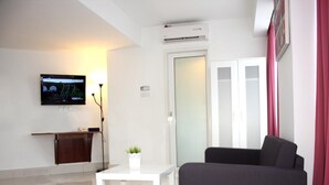 A modern living room with a flat-screen TV mounted on the wall, a small white table with a plant, a dark sofa, and a glass door leading to a bathroom. - V Hotel (Kuala Lumpur)