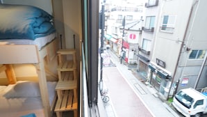 A bunk bed with a blue blanket, a wooden ladder, and a window overlooking a street with buildings and a white van.