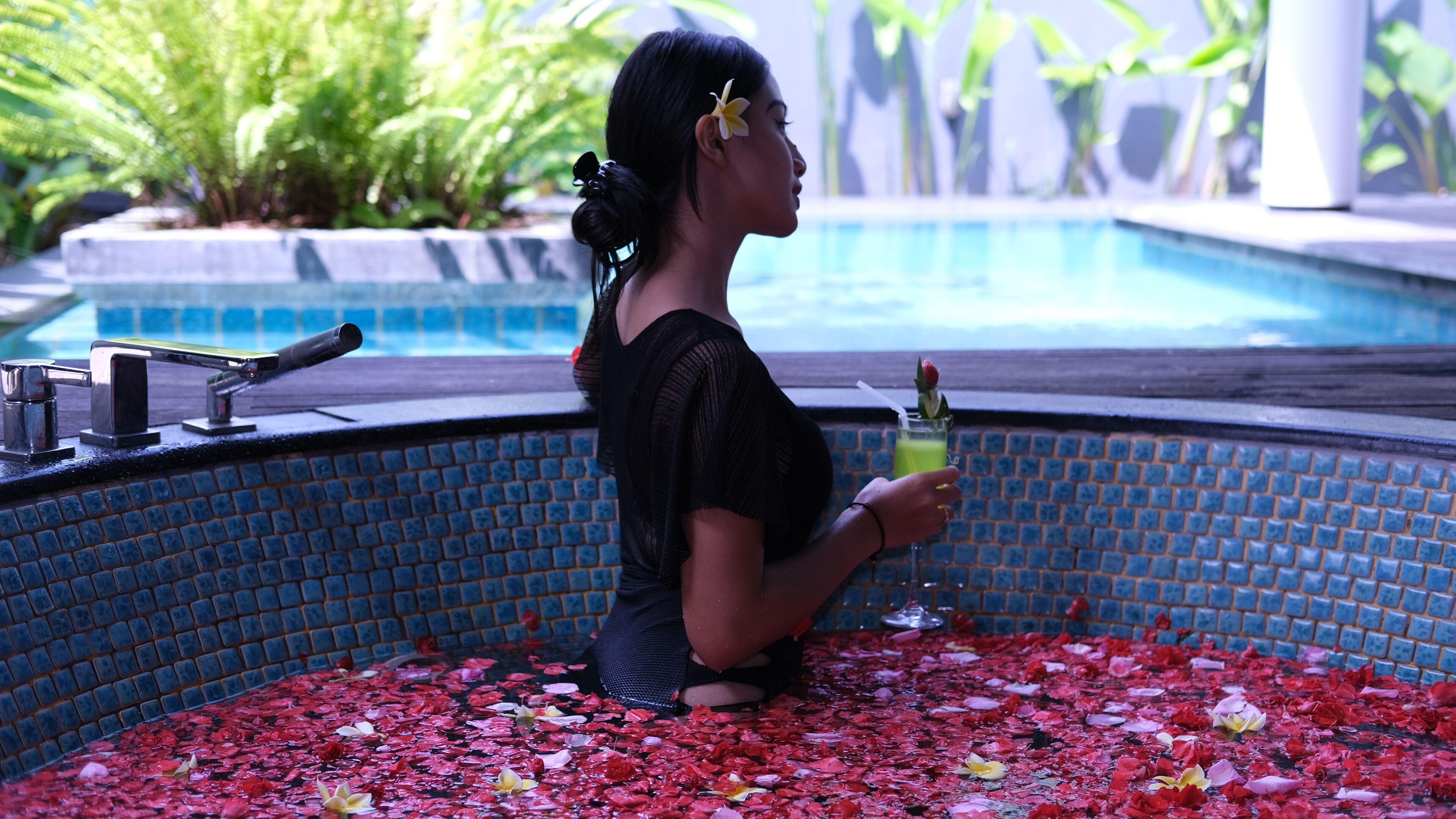 A woman sitting in a pool filled with rose petals, holding a drink.