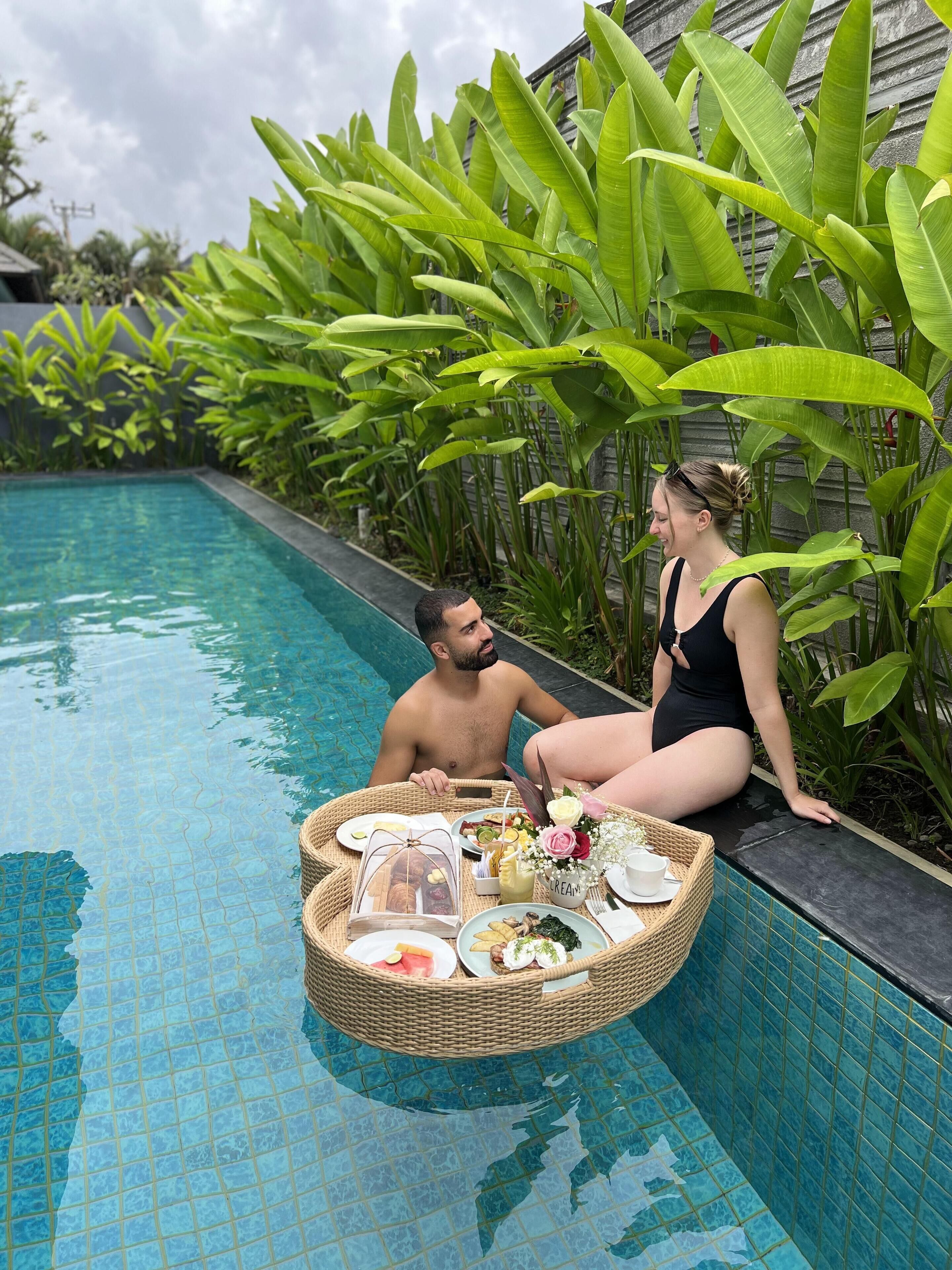 A couple relaxing by a poolside with a wicker table holding food and drinks.