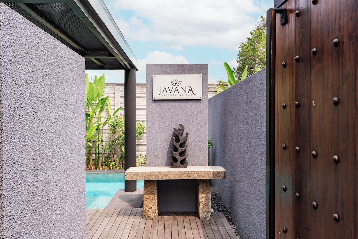Der Eingang einer privaten Villa mit einem Schild, auf dem 'JAVANA PRIVATE VILLAS' steht, einer Steinsitzbank und einer Holztür.