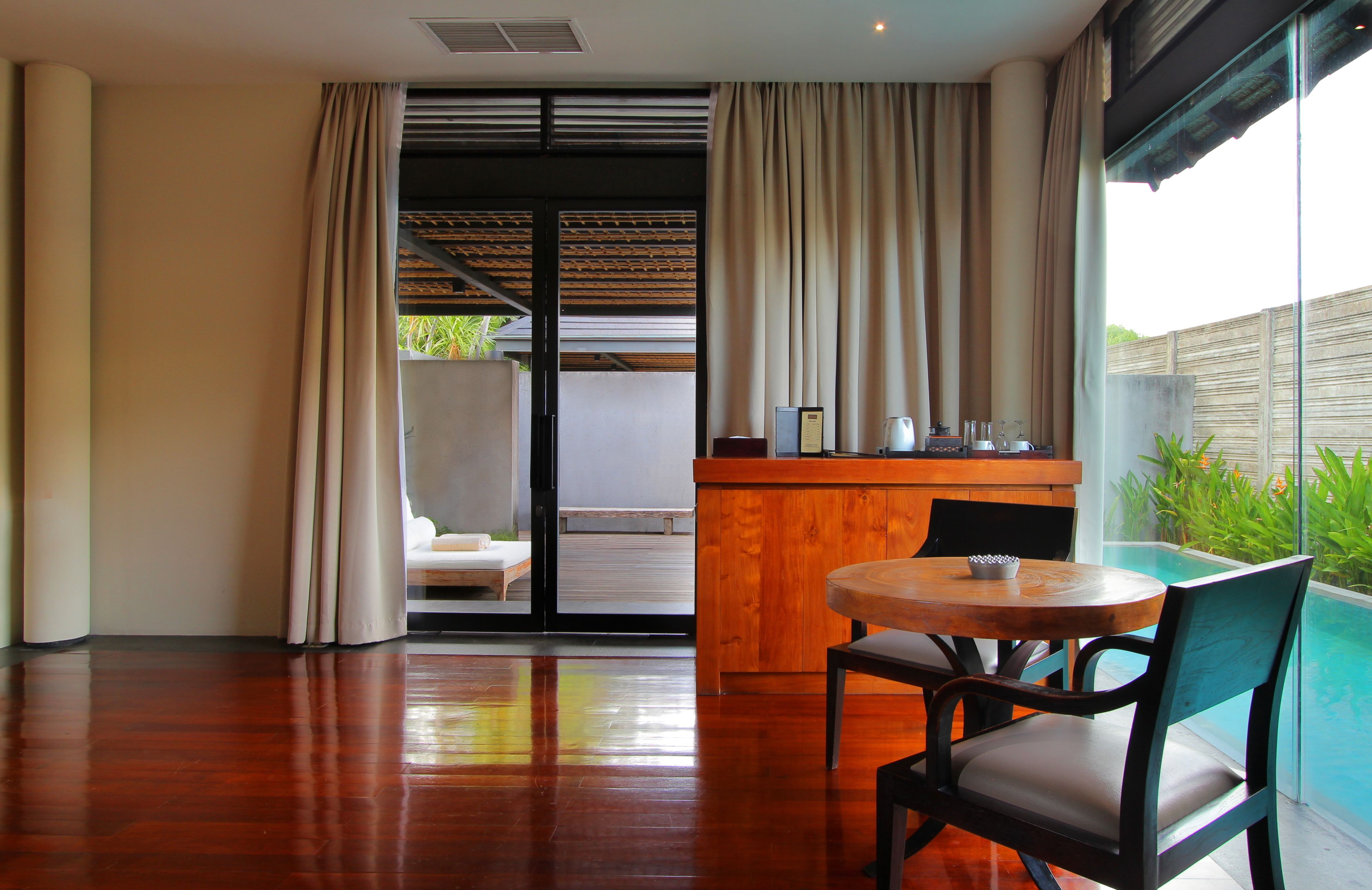 Una habitación moderna con suelo de madera, una mesa redonda con dos sillas, una pequeña barra de madera y un ventanal corredero que da a un balcón con piscina.