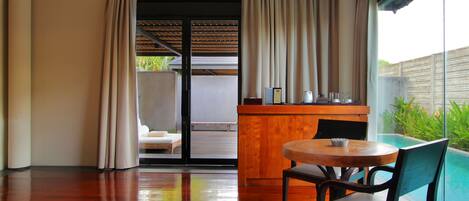 Una habitación moderna con suelo de madera, una mesa redonda con dos sillas, una pequeña barra de madera y un ventanal corredero que da a un balcón con piscina.