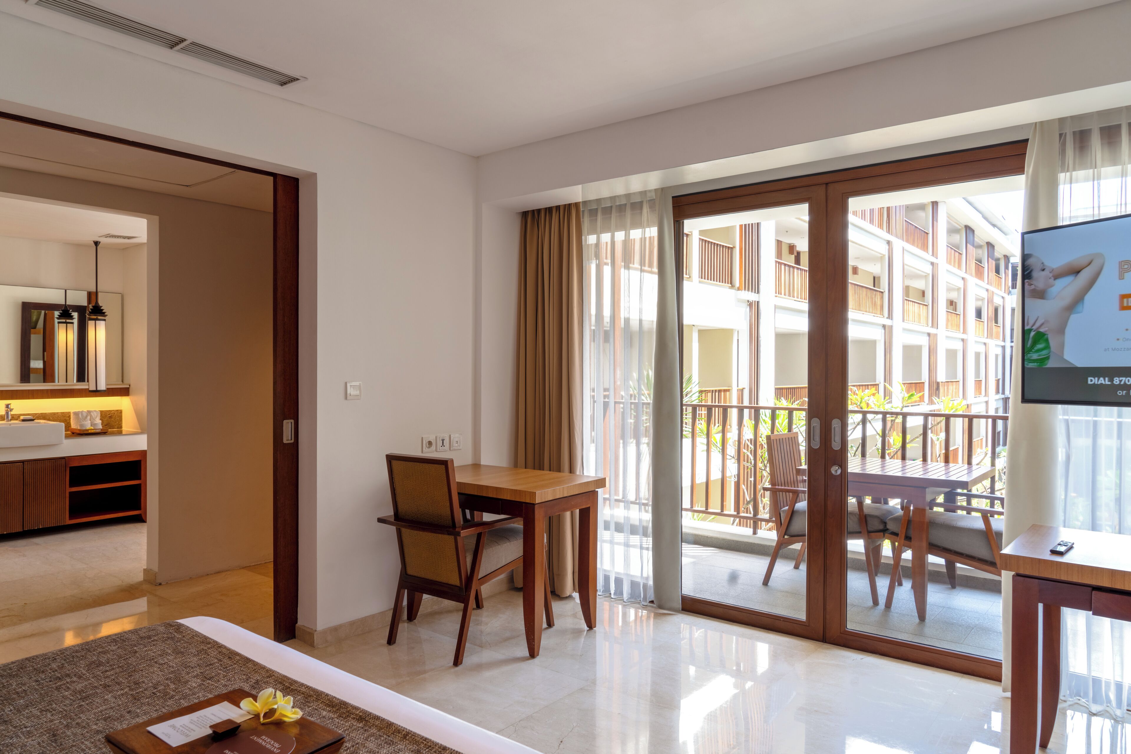 A hotel room with a dining area, a large window with curtains, a flat-screen TV, and a view of the building's exterior.