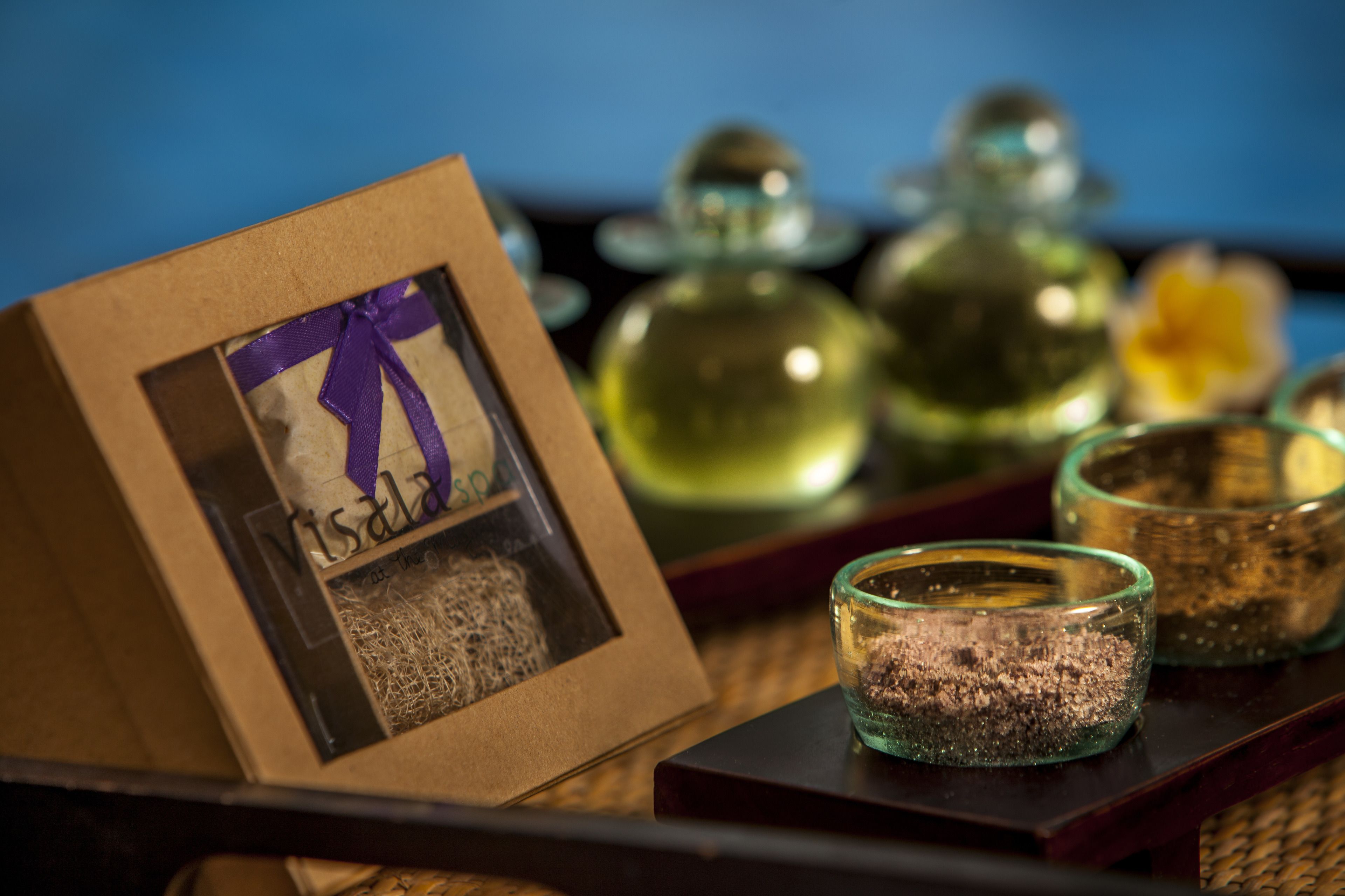 A framed picture with a purple ribbon, a glass candle holder with a textured substance, and two glass containers with a golden liquid.