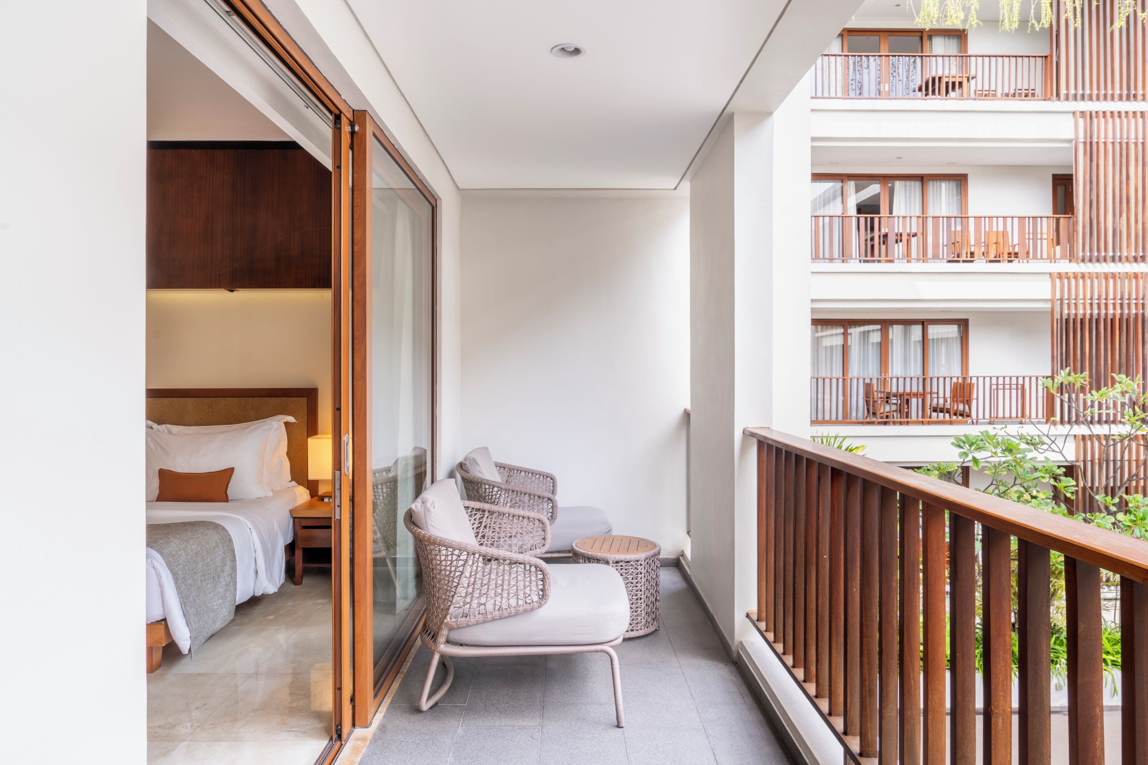Un balcón con mobiliario de mimbre, una cama con ropa de cama blanca y vistas del exterior del edificio.