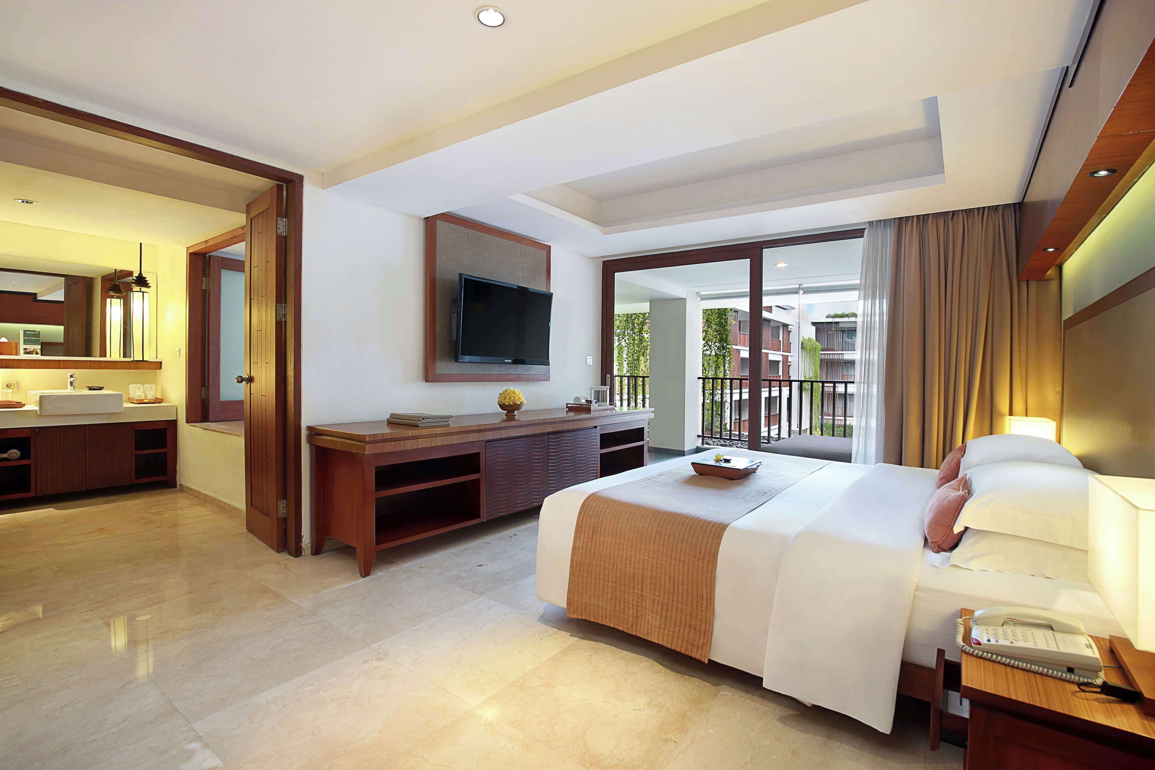 A hotel room with a bed, a desk, a TV, and a balcony with a view of buildings.