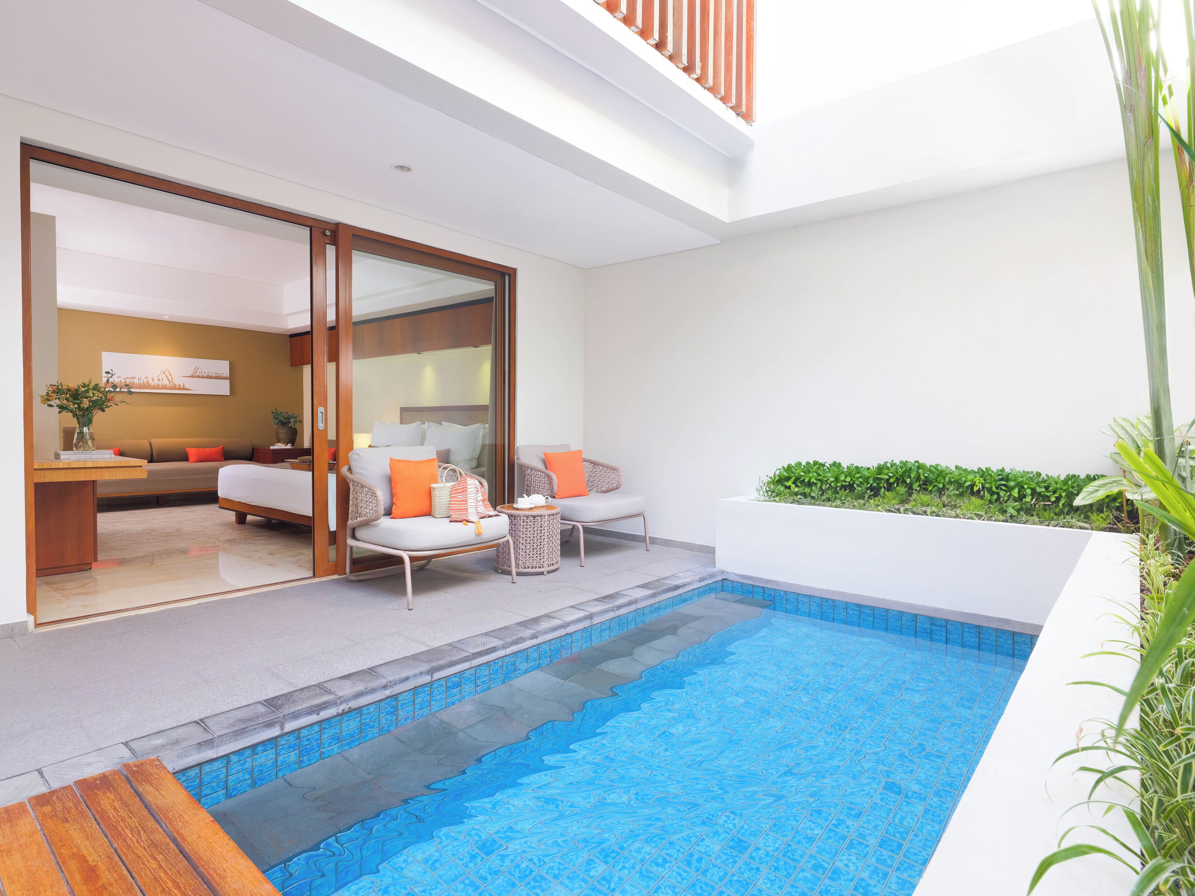 A modern hotel room with a glass door leading to a balcony, a small pool, and a seating area with a sofa and chairs.