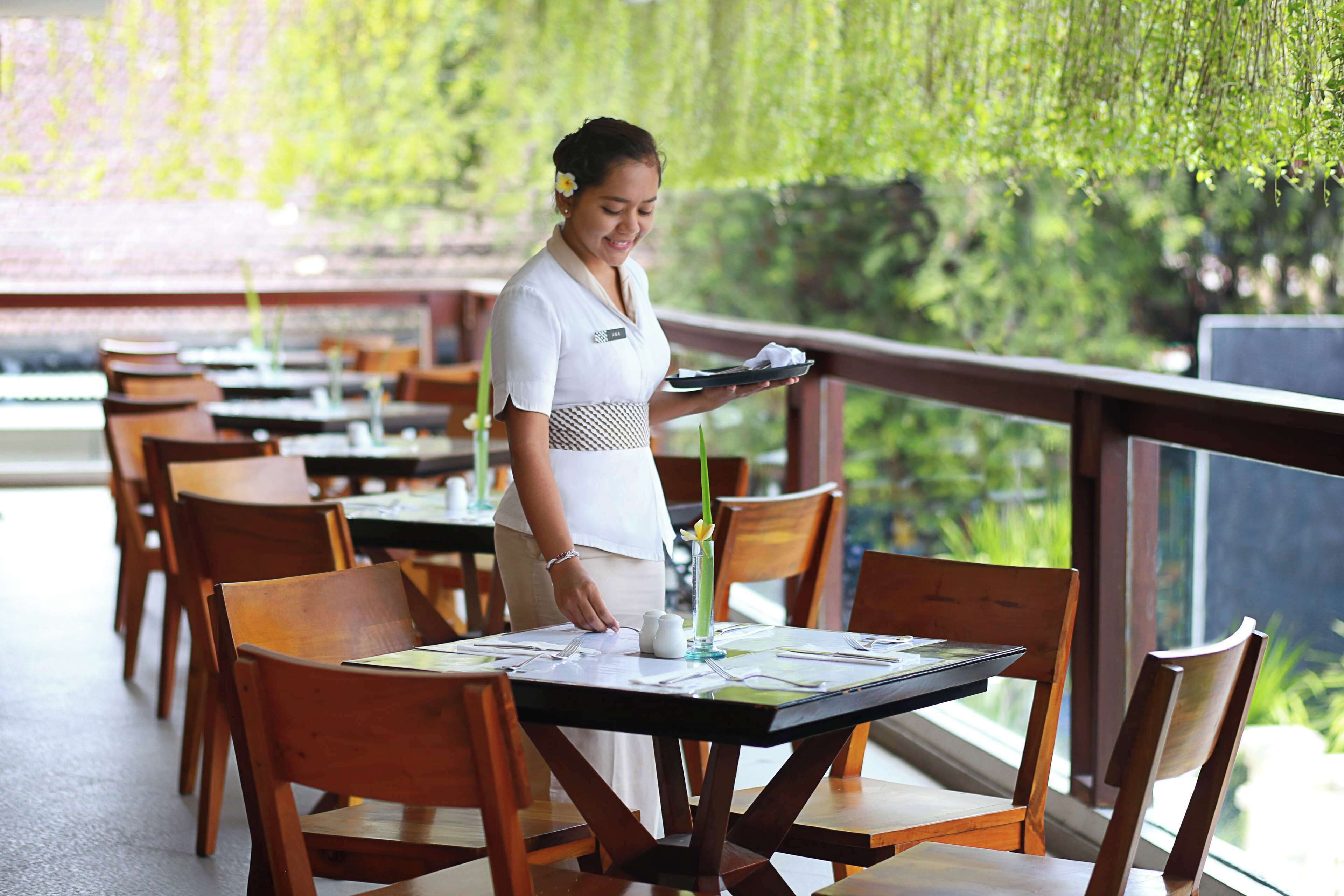 A restaurant with wooden tables and chairs, a waiter holding a tray, and a view of greenery through the windows.