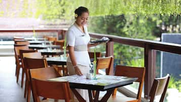 A restaurant with wooden tables and chairs, a waiter holding a tray, and a view of greenery through the windows.