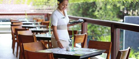 A restaurant with wooden tables and chairs, a waiter holding a tray, and a view of greenery through the windows.