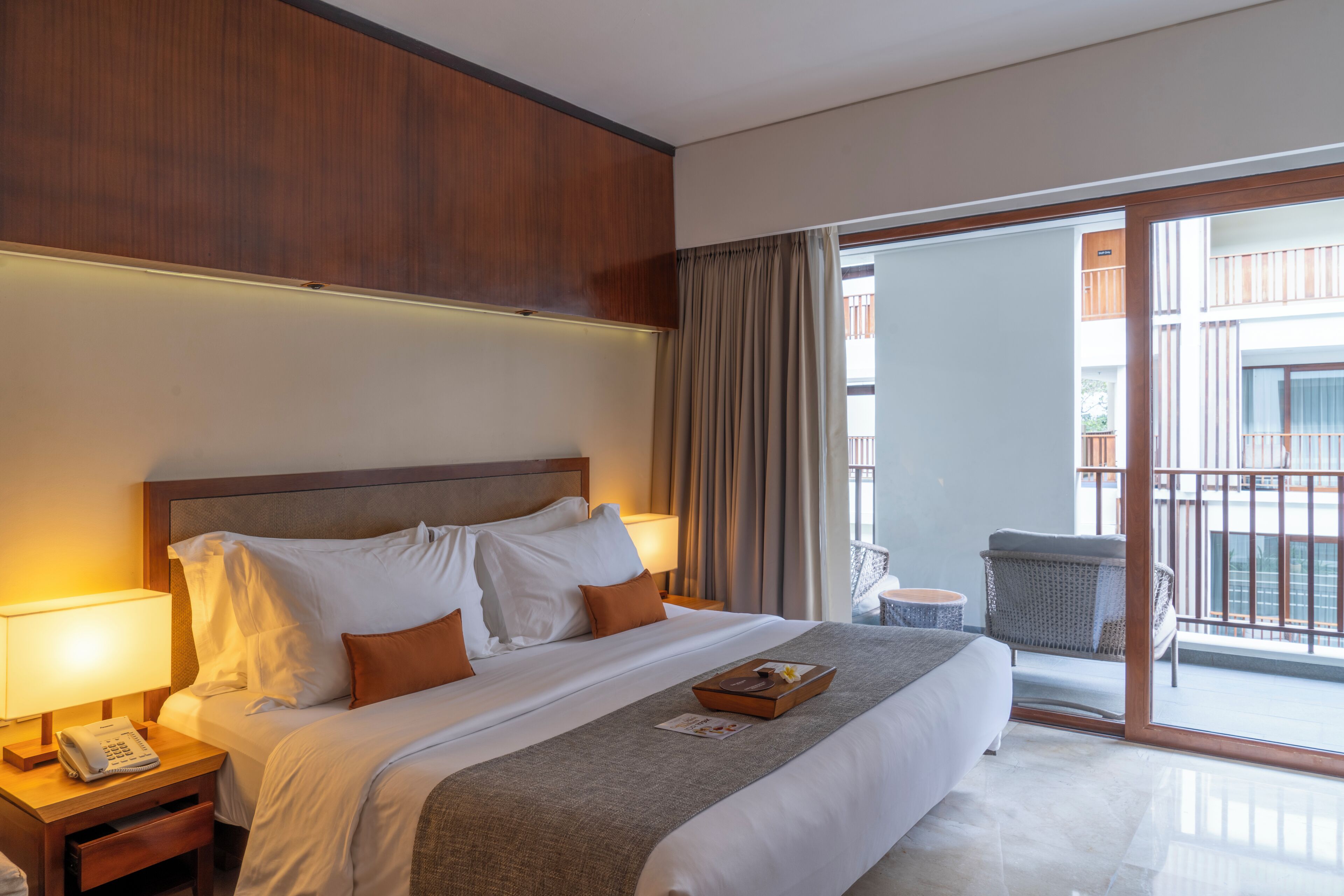 A hotel room with a large bed, a bedside table with a lamp, a small table with a chair, and a view of the outside balcony.