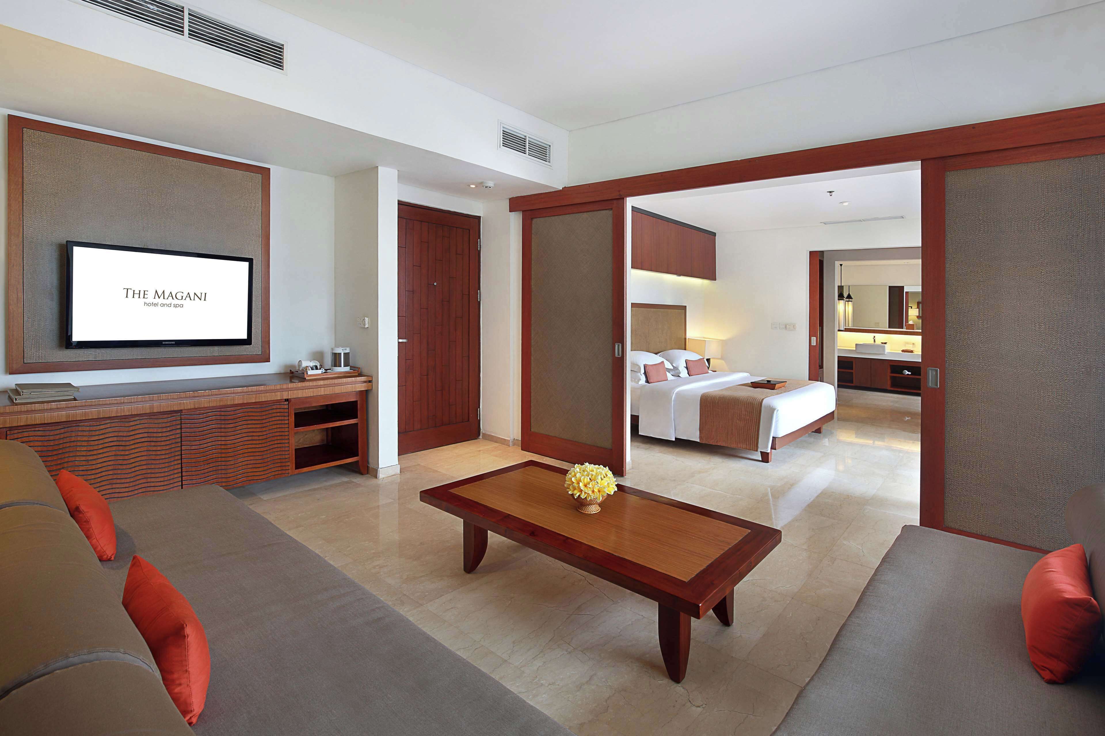 Een hotelkamer met een televisie waarop “THE MAGANI hotel and spa” staat, een bank, een bijzettafeltje met een decoratief object, een bed en een badkamer die door een open deur zichtbaar is.