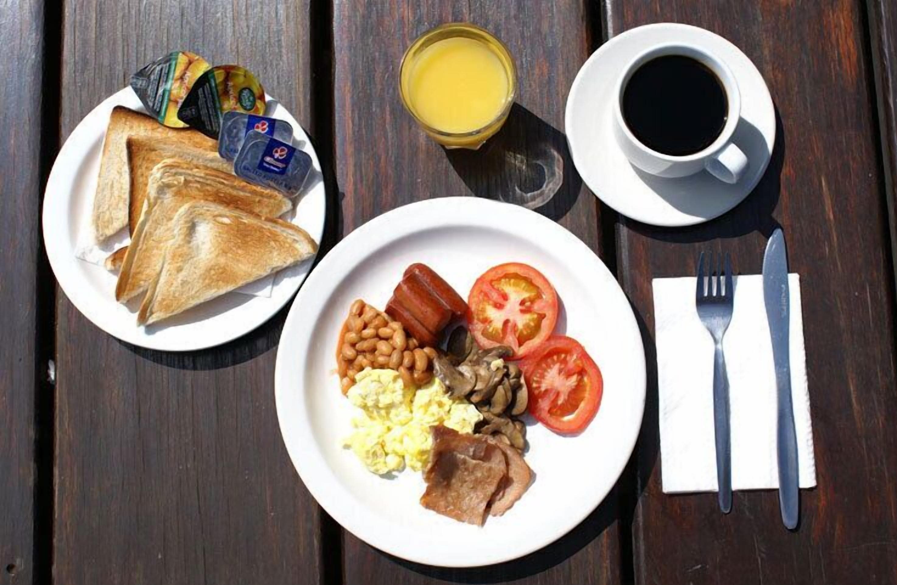 Une table de petit-déjeuner avec des toasts, des œufs, des haricots, des tomates et du bacon sur une assiette, un verre de jus d’orange et une tasse de café sur une table en bois.