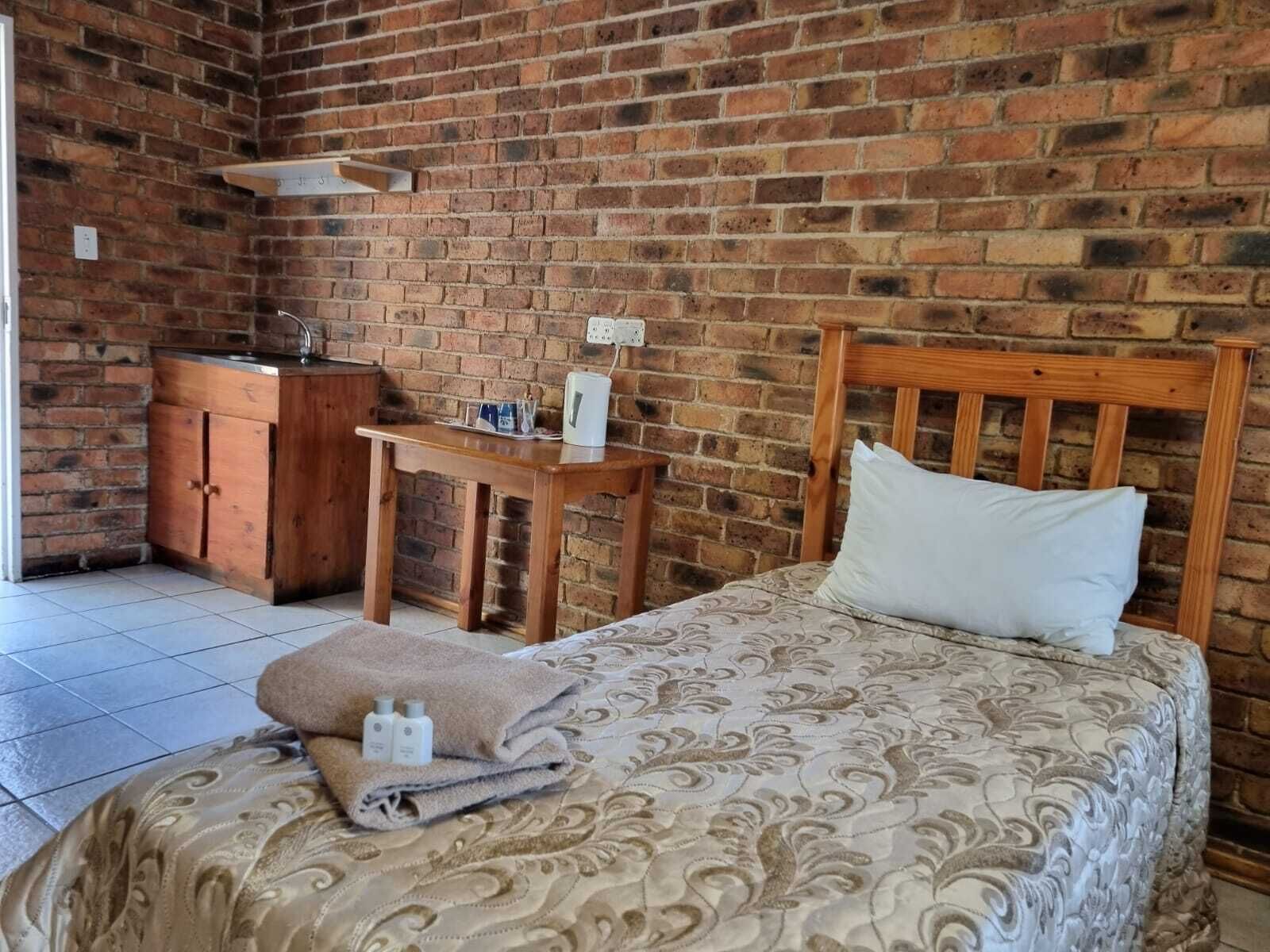 Une pièce avec un mur en briques, un lit en bois recouvert d’une couvre-lit à motifs, une table de chevet en bois, une petite table en bois avec une bouilloire, et une armoire en bois.