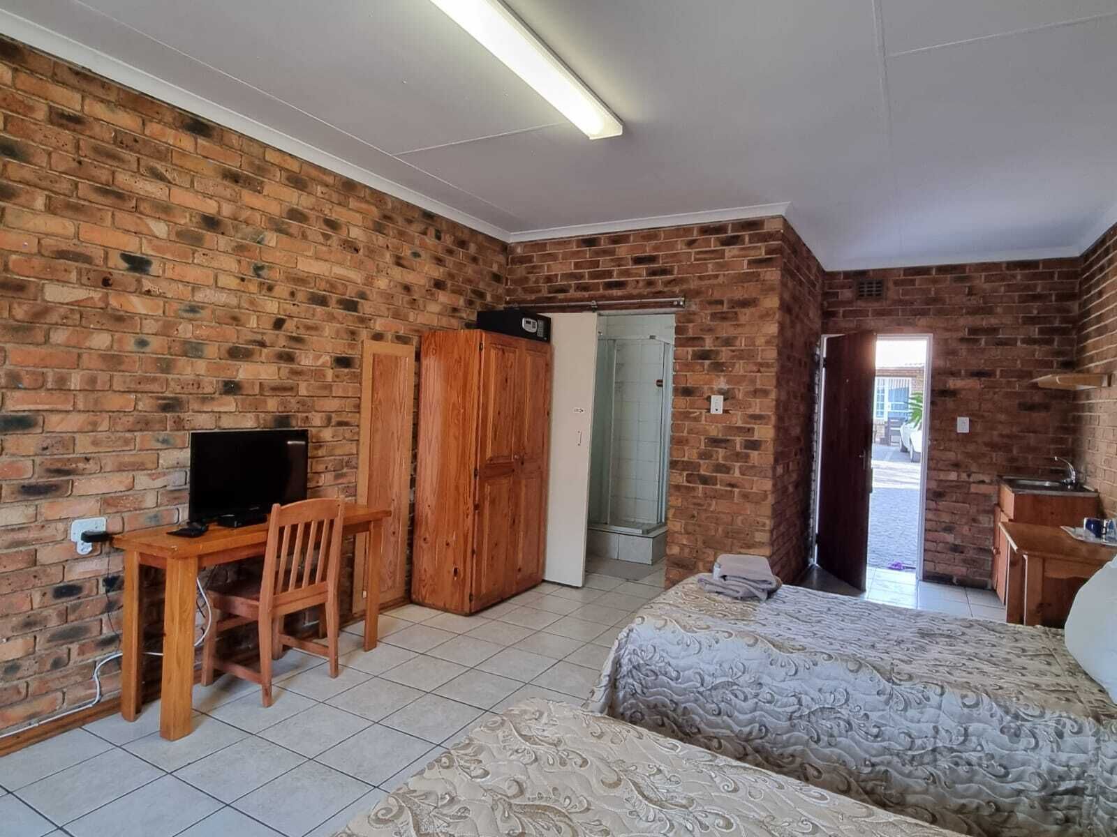 A room with a brick wall, a bed, a desk with a chair, a television, and a bathroom door.