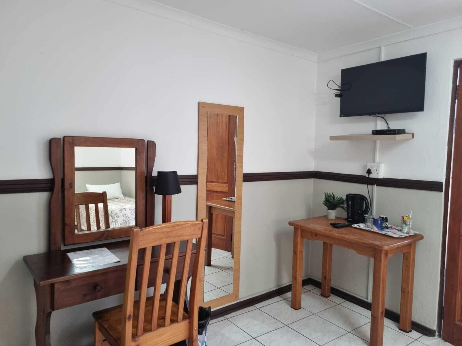 A small, tidy room with a wooden desk, a chair, a mirror, a television mounted on the wall, and a small table with a plant and a kettle.