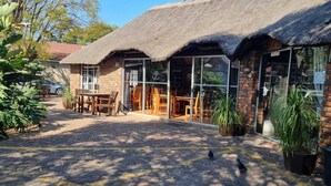 A building with a thatched roof and large windows. There are outdoor tables and chairs, and potted plants around.