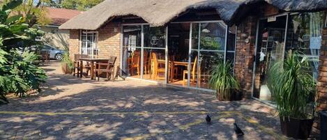 A building with a thatched roof and large windows. There are outdoor tables and chairs, and potted plants around.