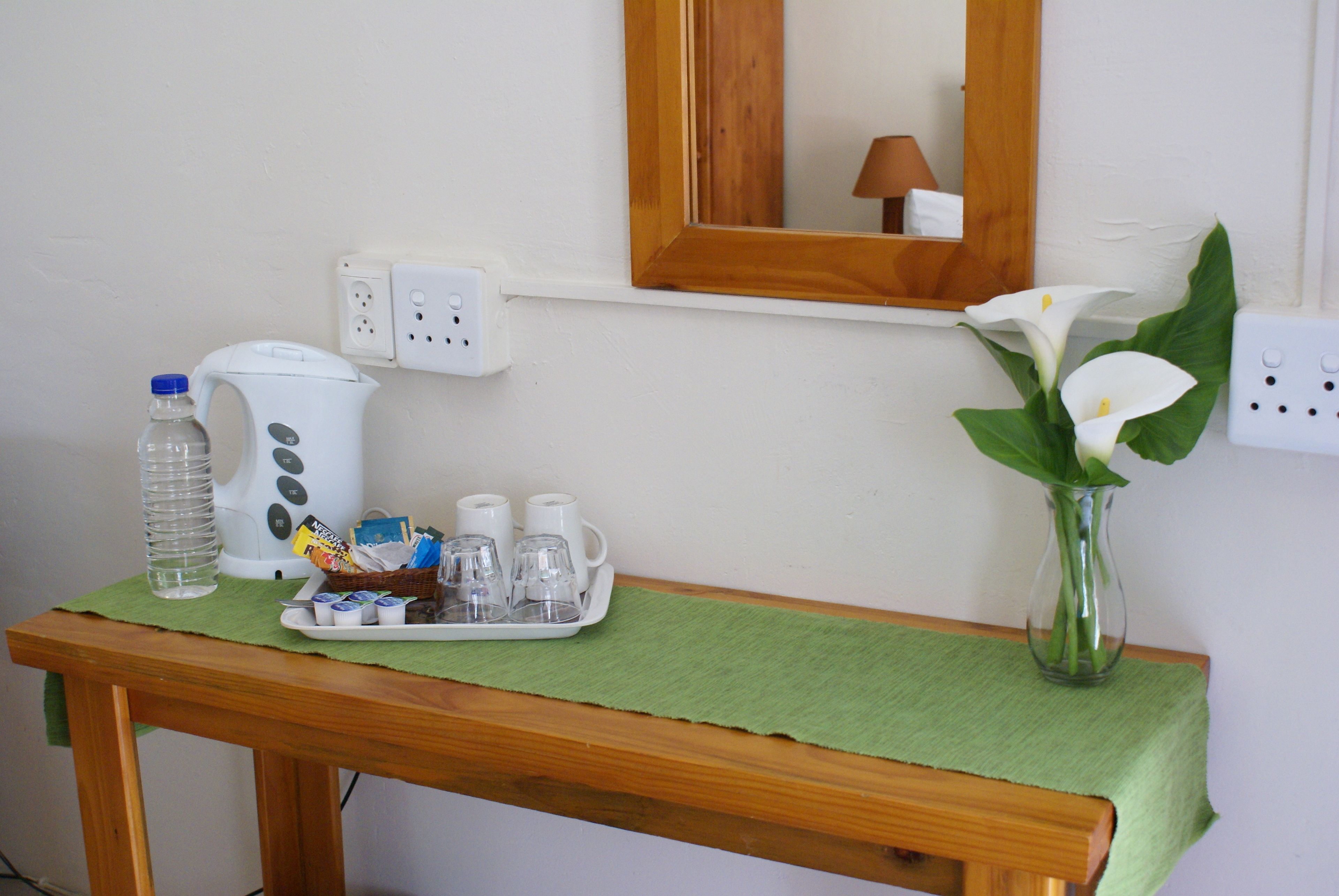 Una mesa de madera con un mantel verde, una botella de agua, una cafetera y una bandeja con tazas y sobres de azúcar. Un jarrón con flores blancas está sobre la mesa.