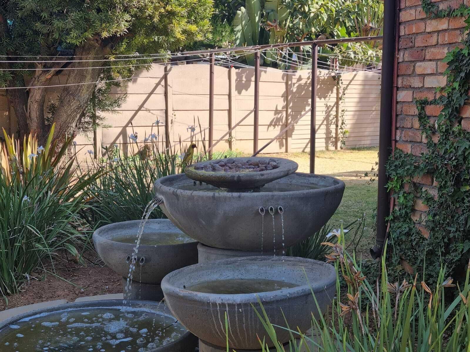 A tiered stone fountain with water flowing from the top tier, surrounded by greenery and a brick wall.