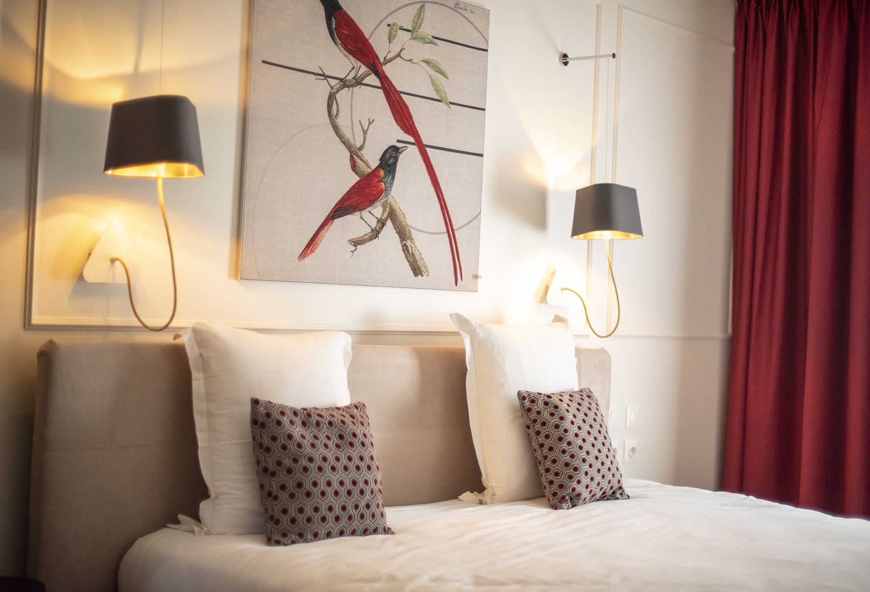 A hotel room with a bed, two patterned pillows, a wall-mounted painting of birds, and two wall-mounted lamps.