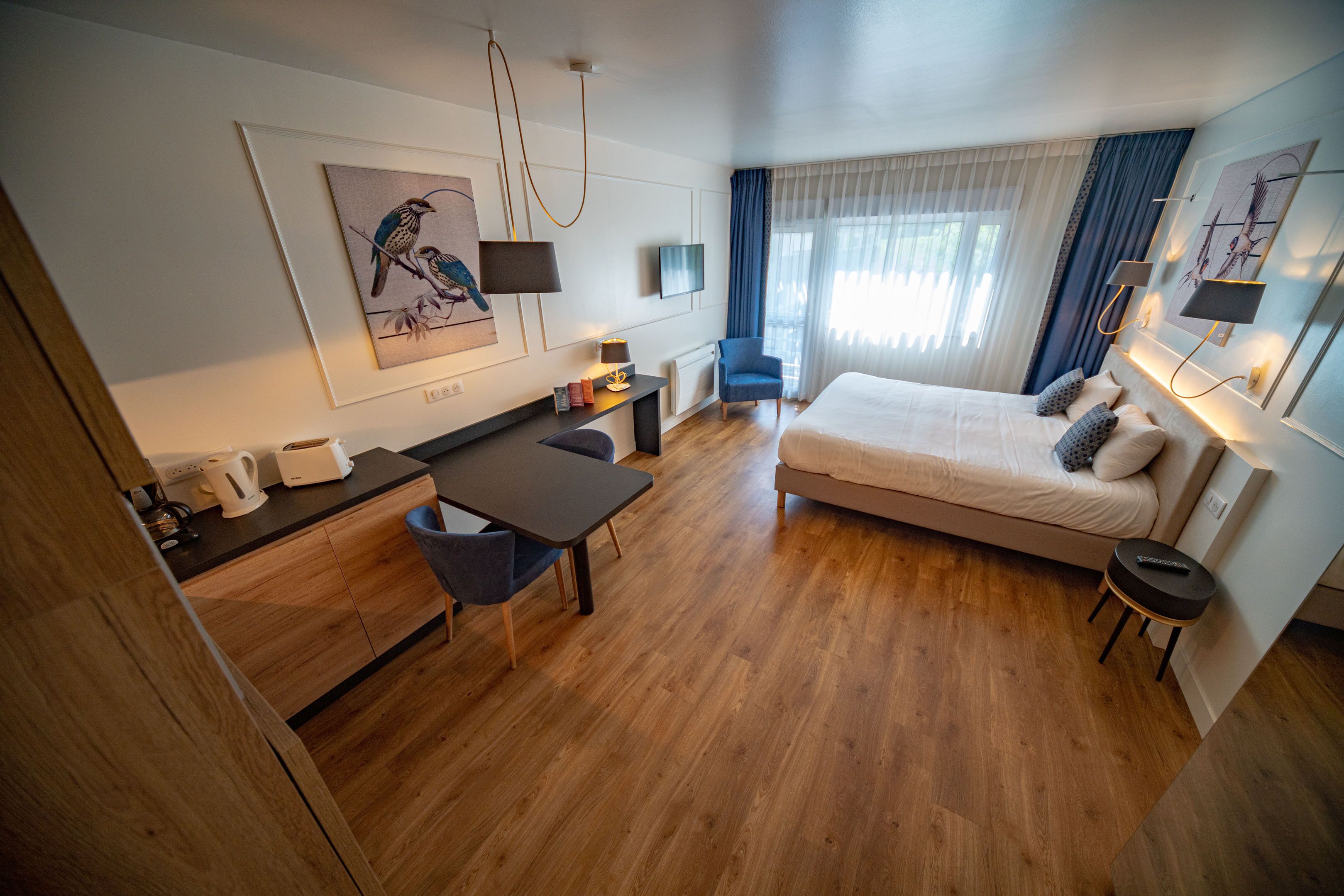 A modern hotel room with a wooden floor, a bed with pillows, a desk with a chair, a coffee maker, and artwork on the walls.