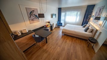 A modern hotel room with a wooden floor, a bed with pillows, a desk with a chair, a coffee maker, and artwork on the walls.