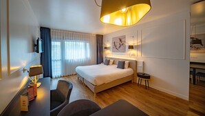 A modern hotel room with a large bed, a desk with a lamp, a chair, and a television.