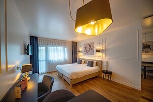 Ein modernes Hotelzimmer mit einem großen Bett, einem Schreibtisch inklusive Lampe, einem Sessel und einem Fernseher.