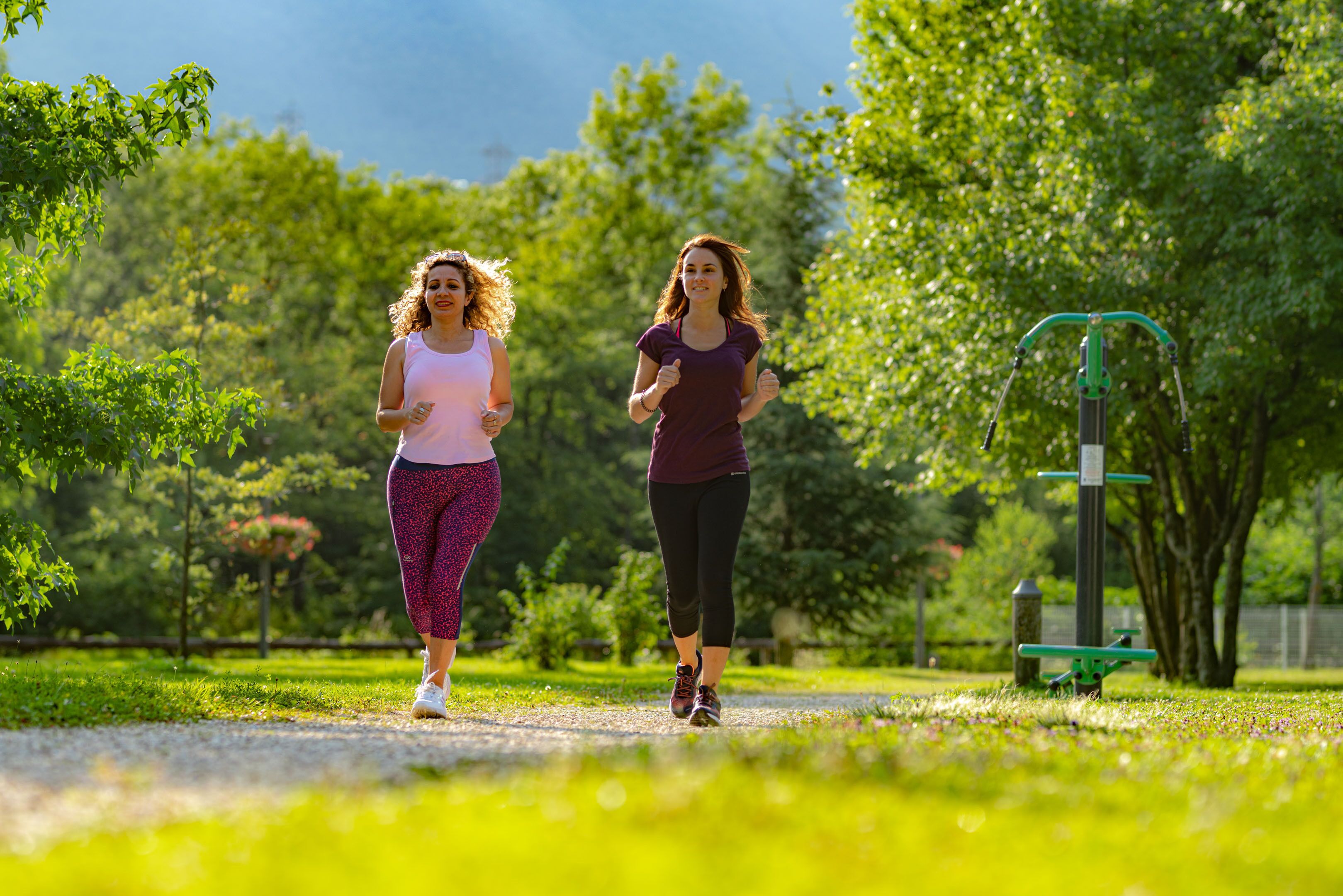 Deux femmes font du jogging sur un sentier dans un parc, avec des arbres et des appareils de musculation en arrière-plan.