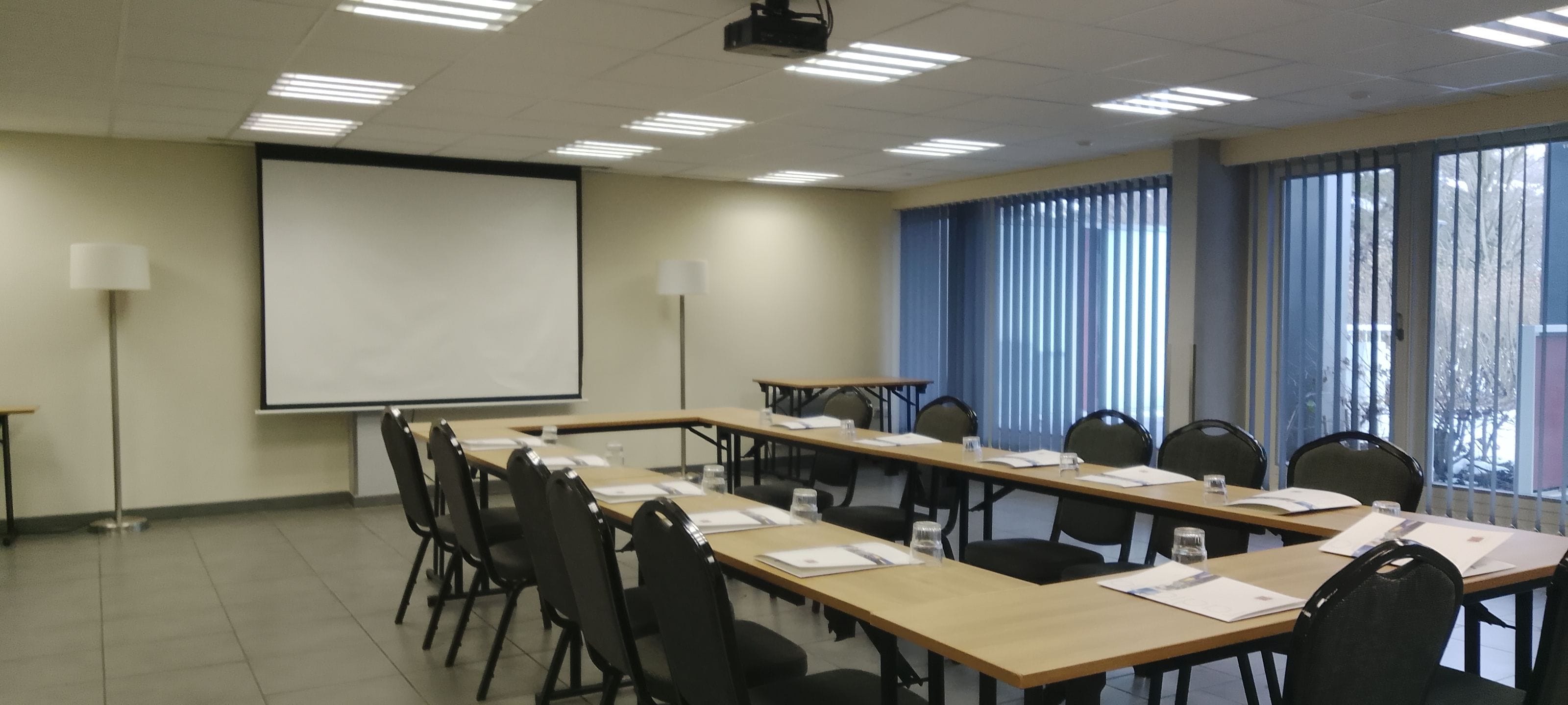 Uma sala de conferências com uma mesa comprida, cadeiras, uma tela de projetor e uma janela com persianas.