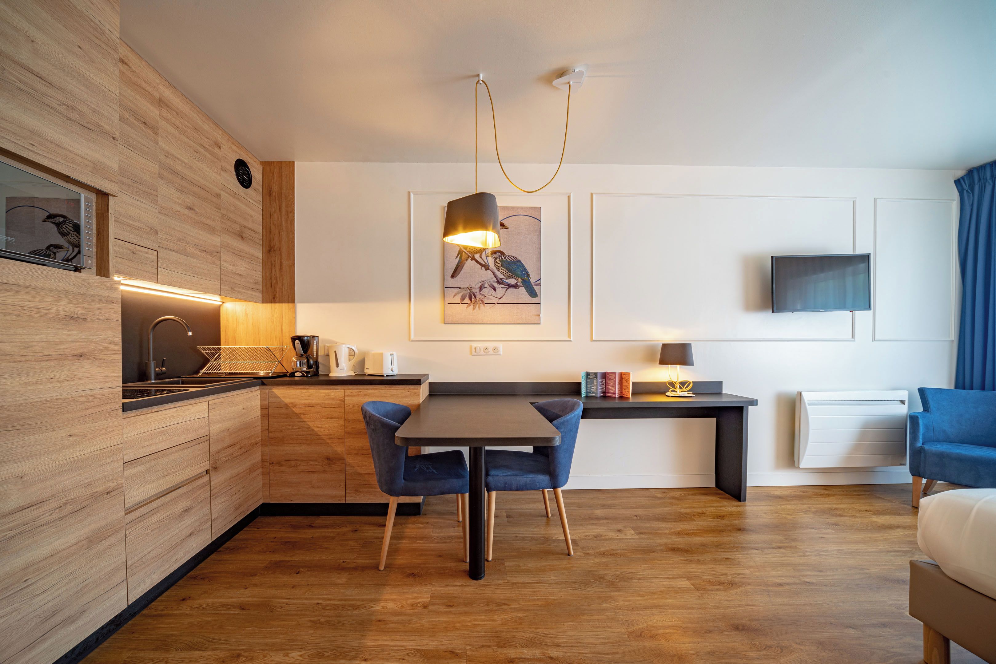 Une cuisine moderne avec des meubles en bois, un coin repas avec une table et des chaises, un téléviseur à écran plat fixé au mur et une lampe fixée au mur.