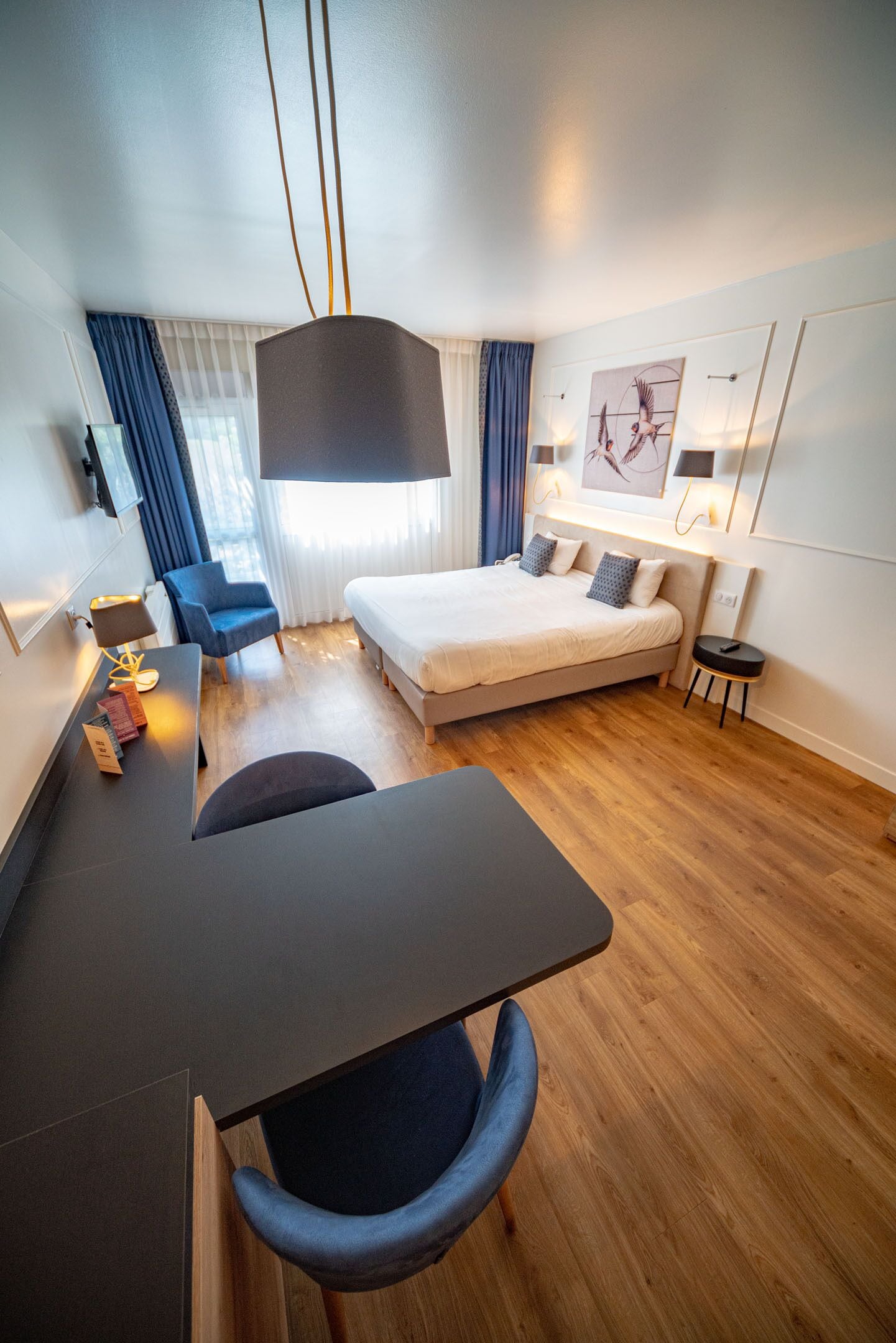 A modern hotel room with a bed, a desk, a chair, a lamp, and a painting on the wall.