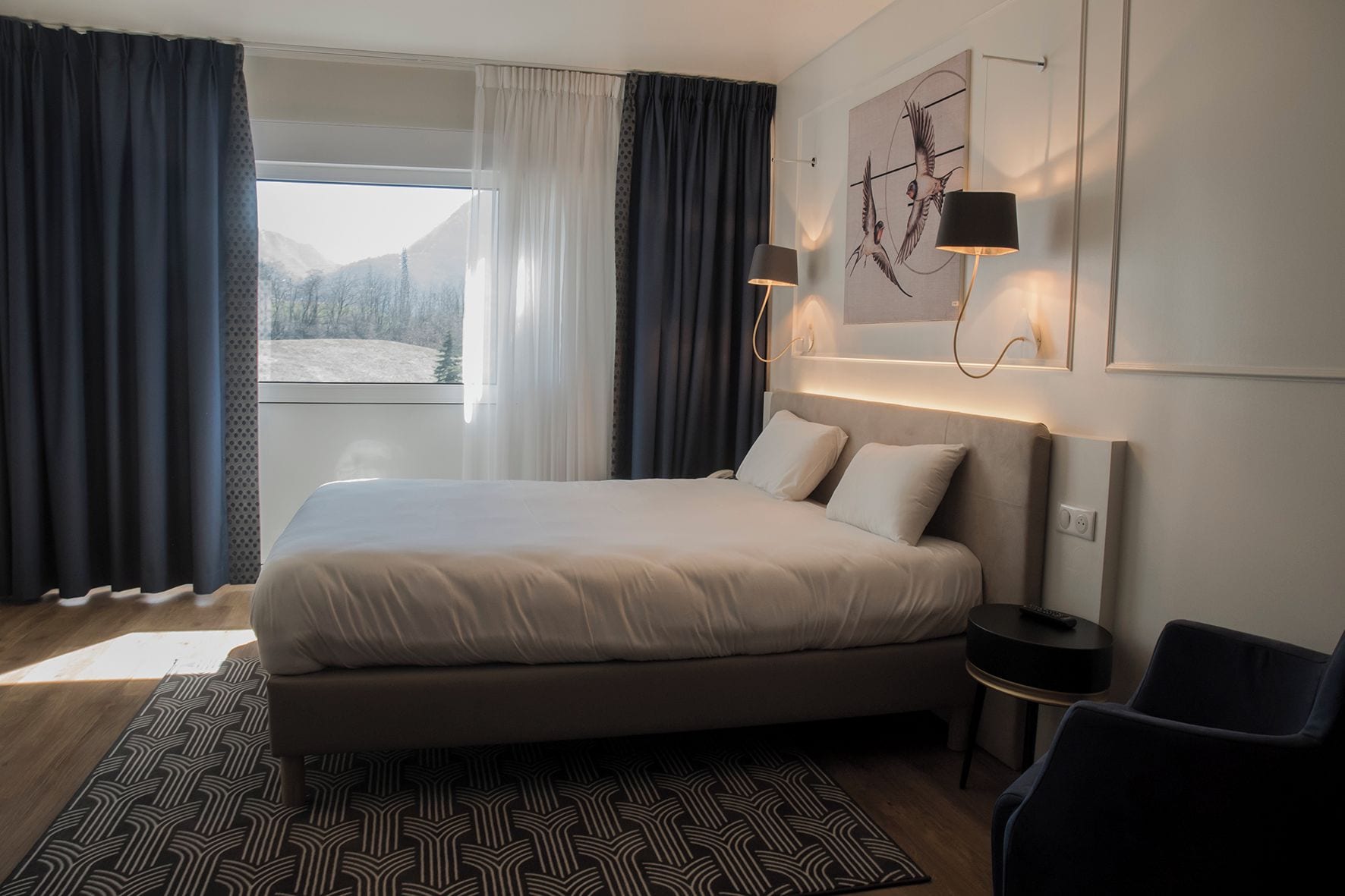 A modern hotel room with a large bed, a bedside table, a chair, and a view of the outdoors.