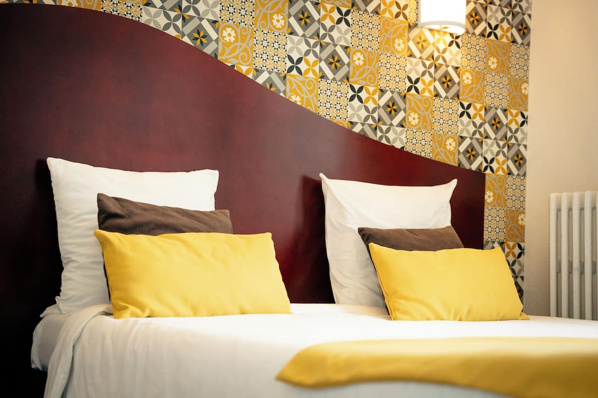 Una cama bien hecha con almohadas blancas y amarillas, un cabecero oscuro y una pared con diseño de fondo.
