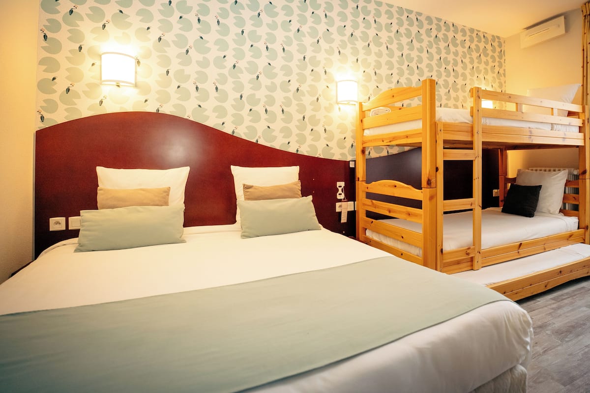Habitación de hotel con dos camas, una con cabecera roja y la otra con literas de madera. Hay luces fijadas a la pared y una ventana con cortinas.